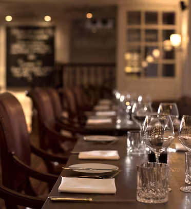 Et varmt opplyst, elegant restaurantinteriør med en rad med polerte trebord dekket med krystallglass, hvite servietter og bestikk. Skinnkledde stoler står langs bordene, og en tavlemeny er synlig i bakgrunnen.