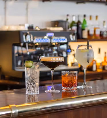En bardisk i tre viser fire drinker: en cocktail med agurkpynt, en espresso martini, en whiskycocktail og en lys drink med is. Bak dem står en espressomaskin og hyller med ulike brennevinsflasker.