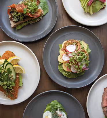 Luftfoto av seks tallerkener med mat på et trebord. Retter inkluderer egg, avokadotoast, en sandwich med grønt, en rett med skiver kjøtt, en panert gjenstand med sitronbåter og en salat med reddiker og grønt.