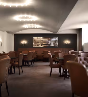 Et svakt opplyst, elegant spiseområde med brune skinnstoler rundt mørke bord. Varme overlys skaper en koselig atmosfære. Et langt speil og lampetter er på veggene, og sollys filtreres gjennom et vindu til høyre.