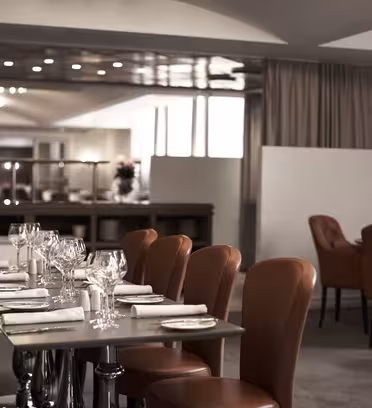 Et elegant restaurantinteriør med pent dekkede bord, med brune skinnstoler og hvite servietter. Myk belysning skaper en varm atmosfære. Bakgrunnen viser store gardiner og speil, noe som forbedrer plassen.