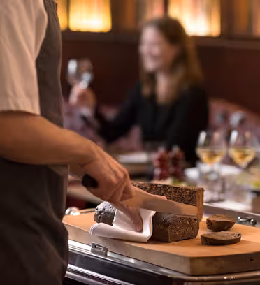 En kokk i hvit skjorte og forkle skjærer mørkt brød på en treplate. En kvinne i bakgrunnen, som sitter ved et bord med vinglass, smiler og hever glasset i et svakt opplyst restaurantmiljø.