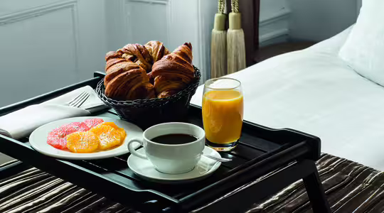 Et frokostbrett på en seng har en svart kurv med croissanter, en tallerken med sitrusfrukter i skiver, en kopp kaffe og et glass appelsinjuice. Oppsettet er på et trebrett med brunt stoff.